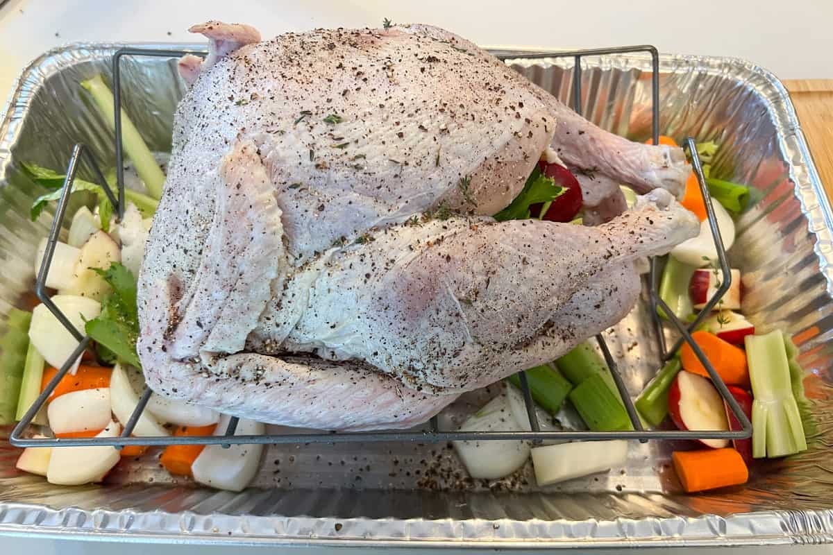 seasoned turkey on a V shaped rack placed inside an aluminum foil pan with veggies all around 