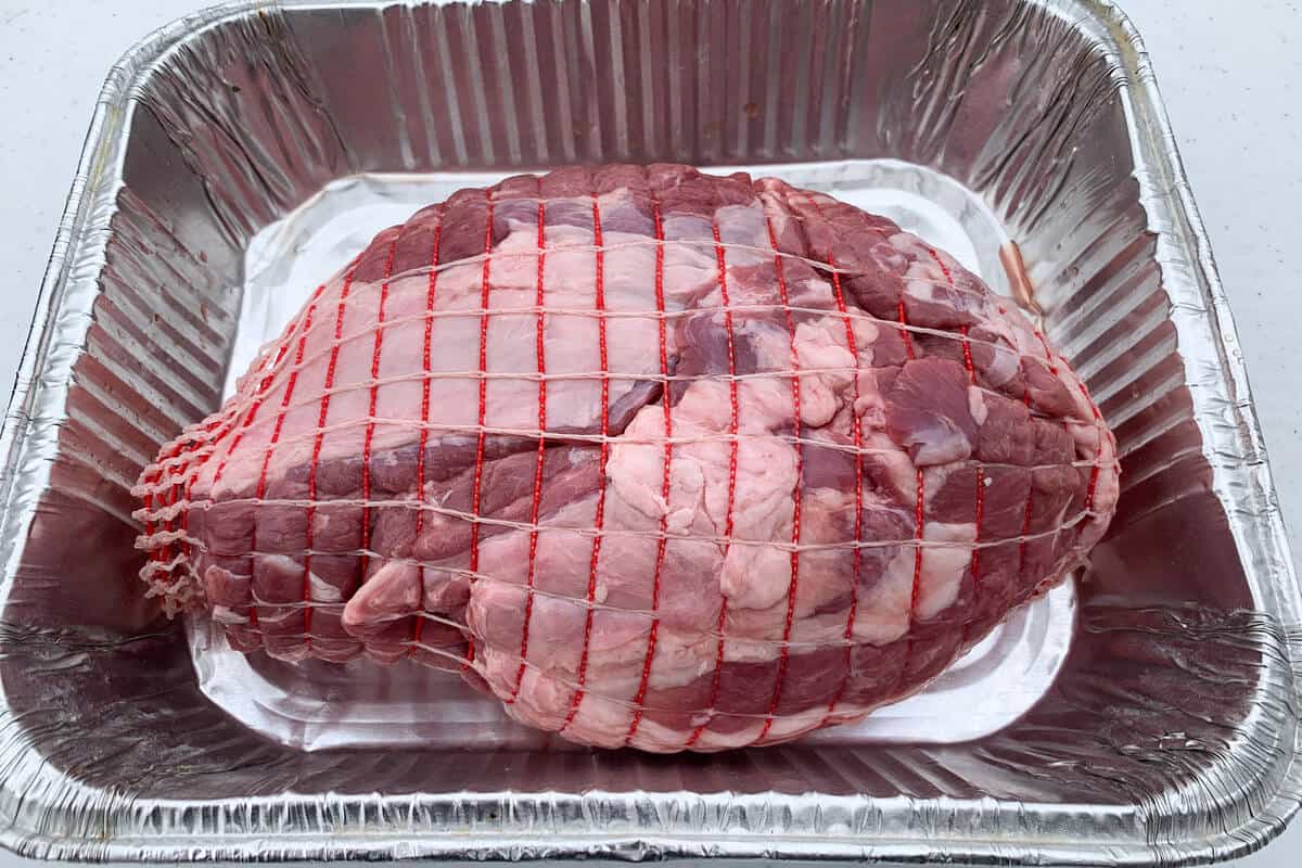 a raw leg of lamb with a net around it in an aluminum foil pan