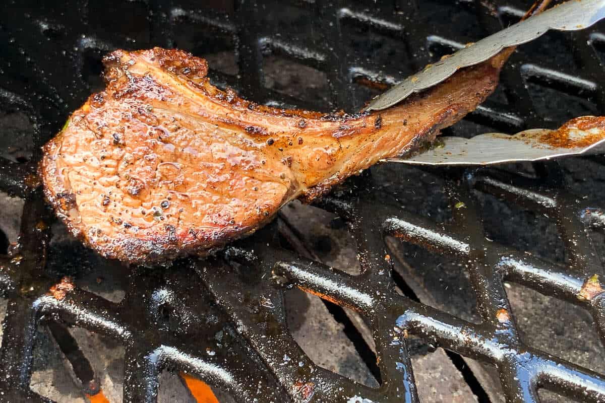 a lamb chop being held with tongs over the flames of a pellet grill