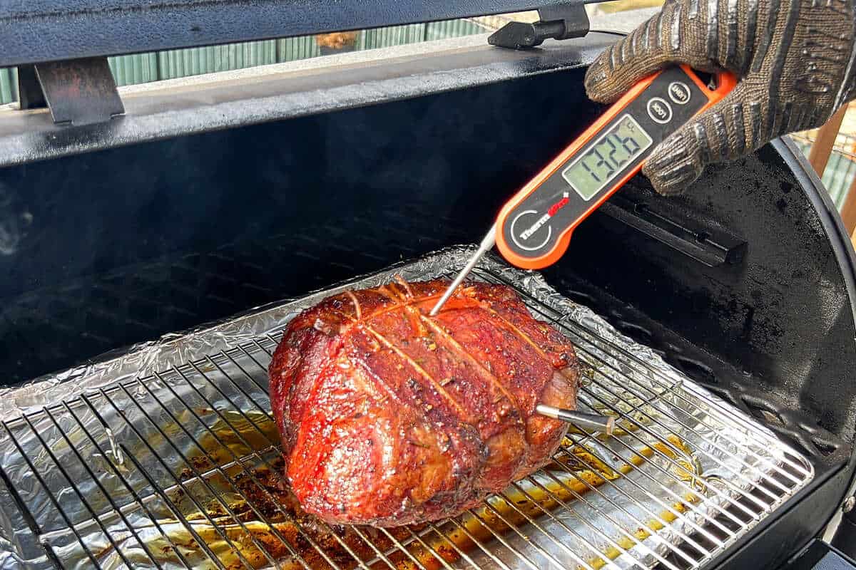 a leg of lamb on a wire rack smoking in a pellet grill with an instant read thermometer reading 132.6 degrees