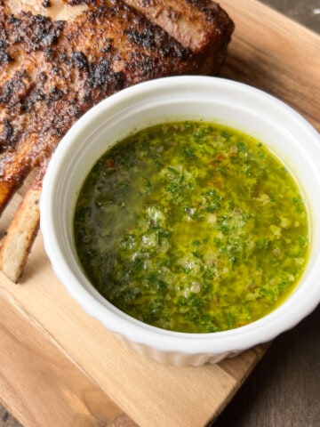 chimichurri sauce in a white bowl on a wooden cutting board with a rack of lamb in the background