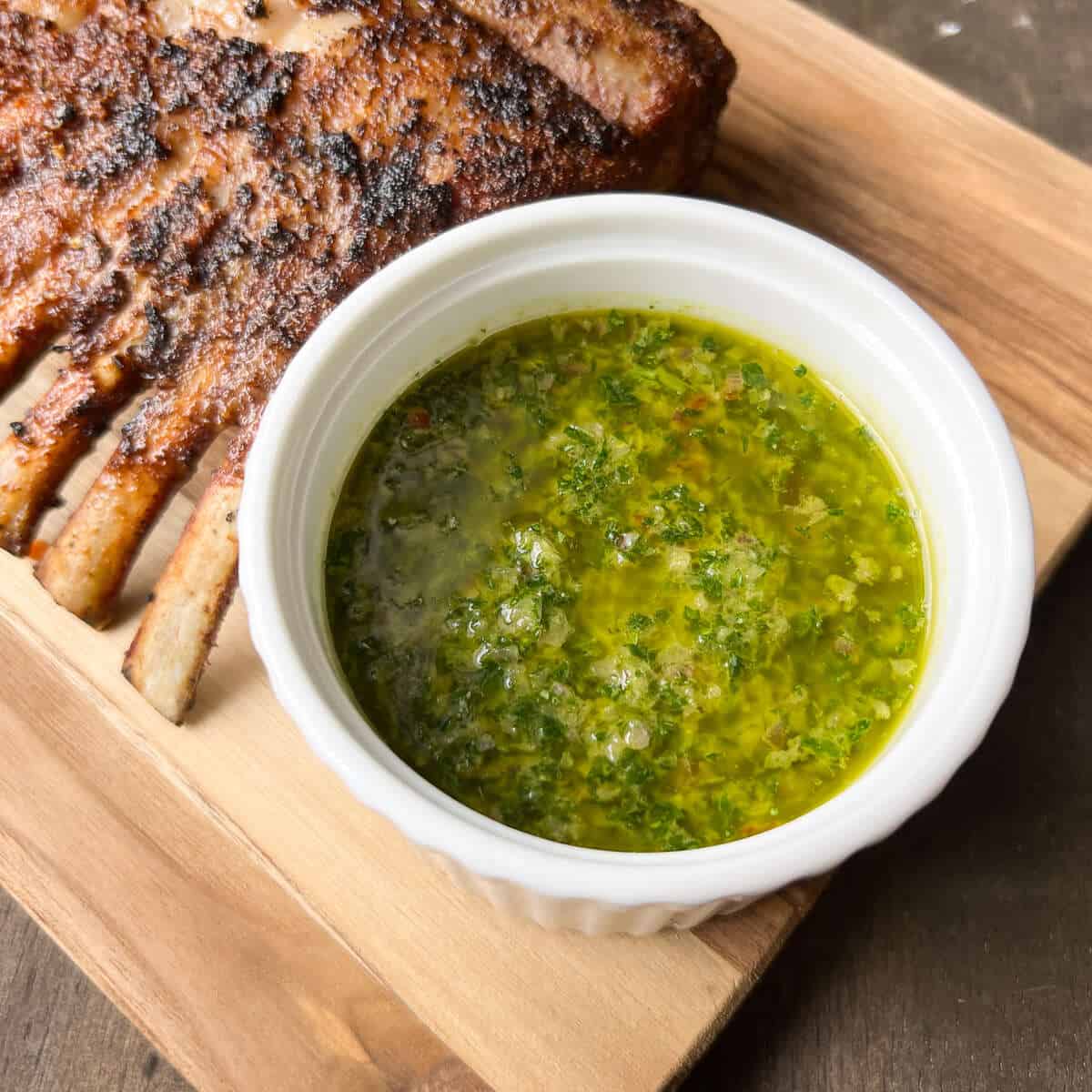 chimichurri sauce in a white bowl on a wooden cutting board with a rack of lamb in the background