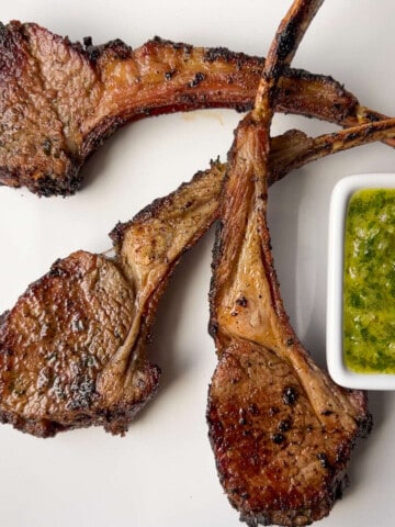 smoked lamb chops on a white plate with a white square dish of chimichurri sauce