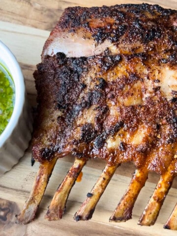 smoked rack of lamb on a wooden cutting board with a white bowl of chimichurri sauce next to it