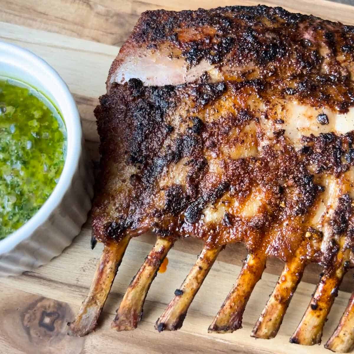 smoked rack of lamb on a wooden cutting board with a white bowl of chimichurri sauce next to it