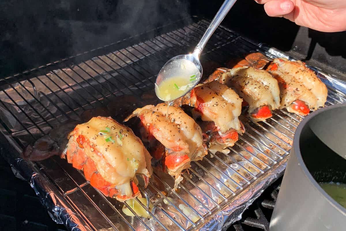 spooning lemon garlic butter over lobster tails on a wire rack over a baking sheet on the grates of a pellet grill