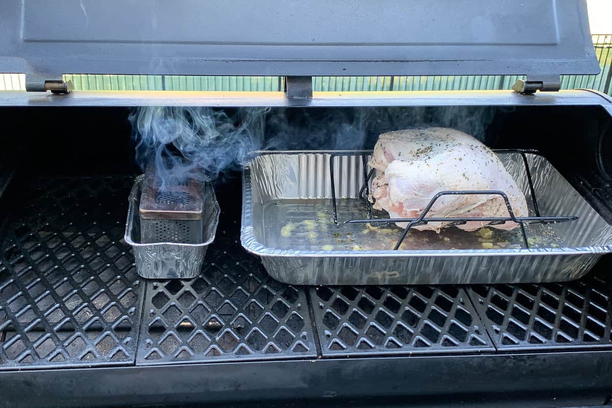 turkey breast on a v shaped rack inside an aluminum foil pan on the grates of a pit boss pellet grill with a smoker box to the left
