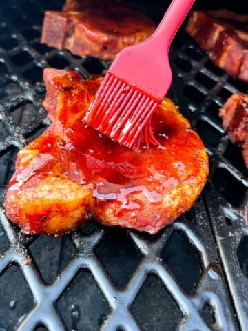 pork chop on the grates of a pit boss pellet grill being sauced with a silicone basting brush