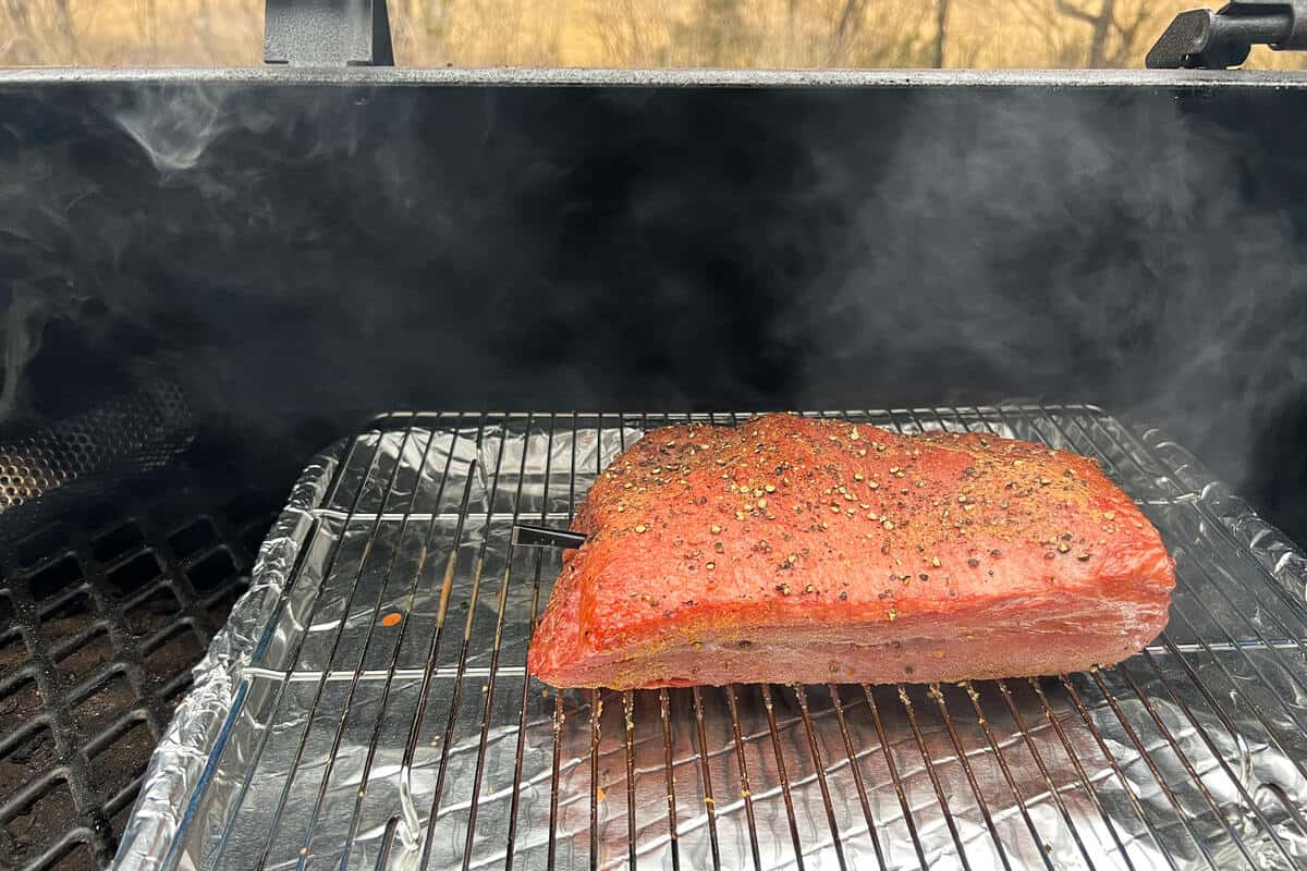 a corned beef brisket on top of a wire rack over an aluminum foil lined baking sheet on the grates of a pit boss pellet grill with a smoker tube on the left side