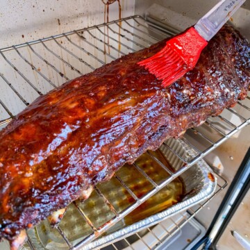 baby back ribs on the grates of a masterbuilt electric smoker with BBQ sauce being brushed on