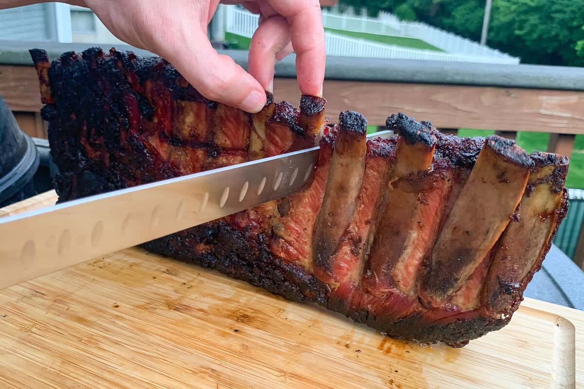 a rack of spareribs held standing up on a cutting board with a meat slicing knife cutting between two of the bones