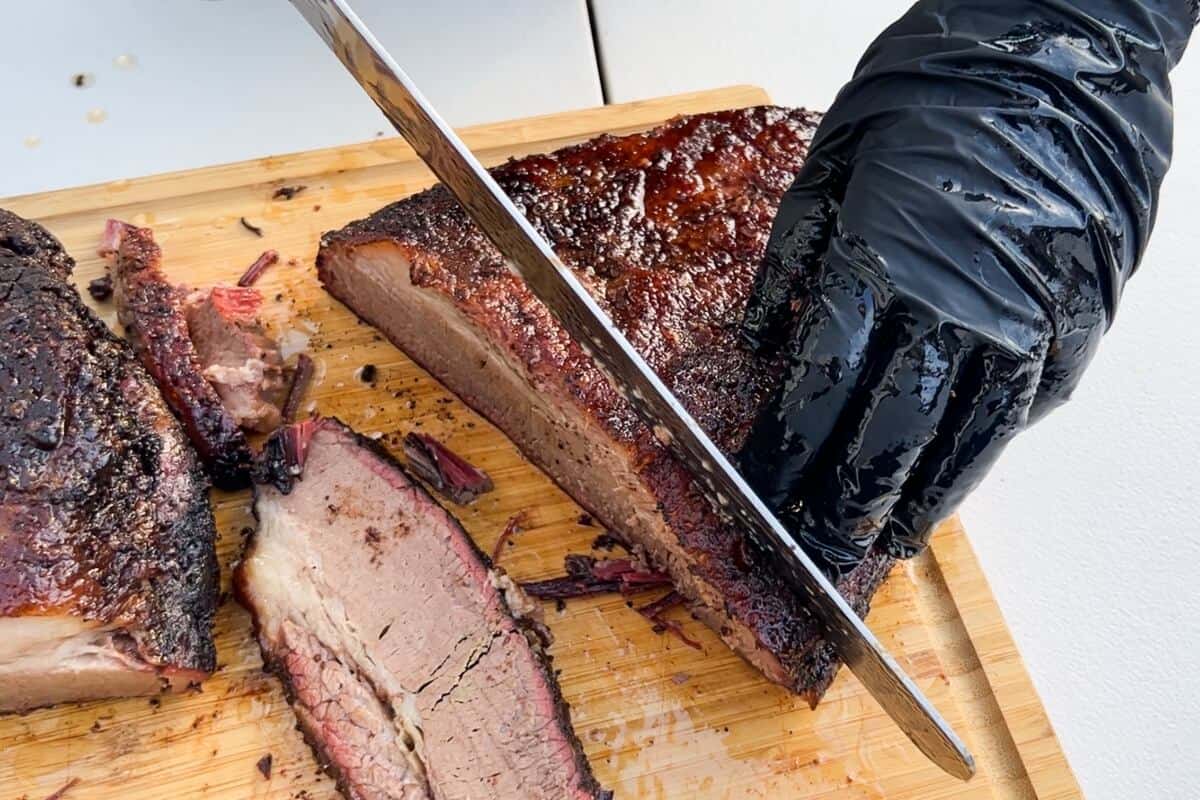 black vinyl gloved hand slicing a brisket with a meat slicing knife on a cutting board