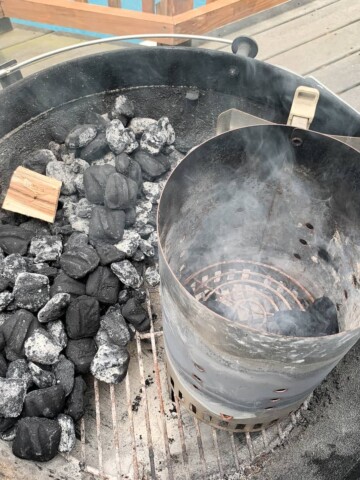 charcoal grill with briquettes piled on one side and a wood chunk on top and a charcoal chimney next to it with lit briquettes inside