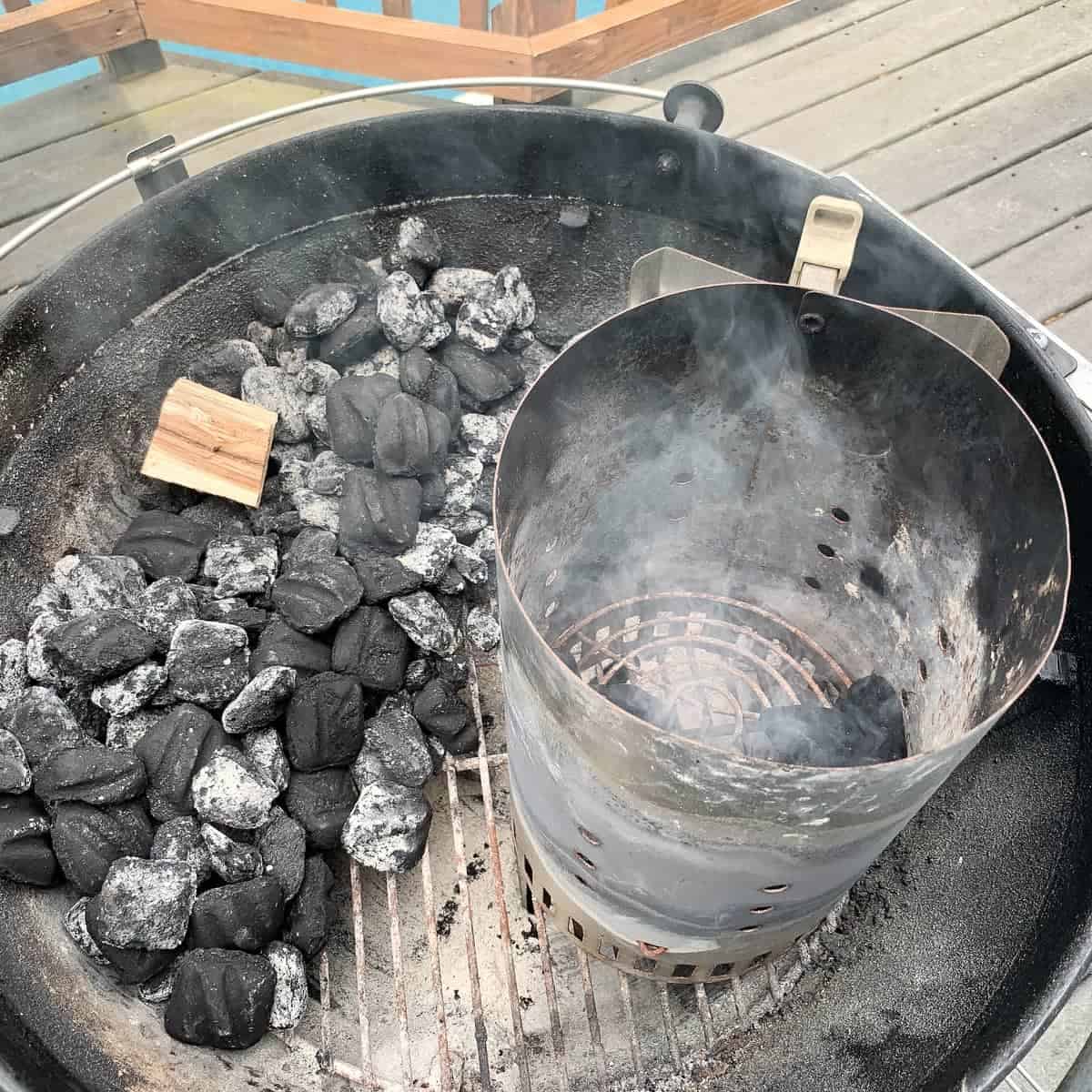 charcoal grill with briquettes piled on one side and a wood chunk on top and a charcoal chimney next to it with lit briquettes inside