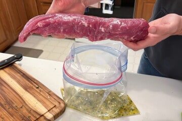 a raw pork tenderloin being held over a gallon size bag filled with marinade