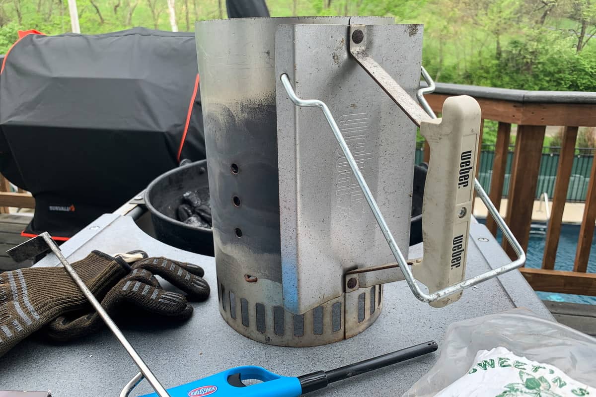 charcoal chimney, heatproof gloves, and a wand lighter on the table of a charcoal grill