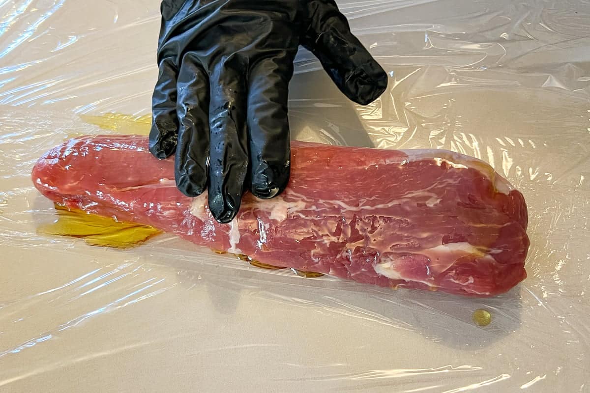 a gloved hand rubbing oil on a pork tenderloin