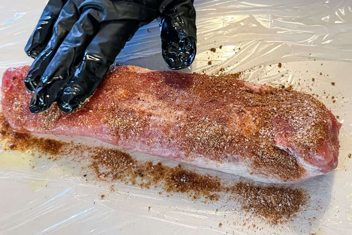 a gloved hand spreading seasoning on a pork tenderloin