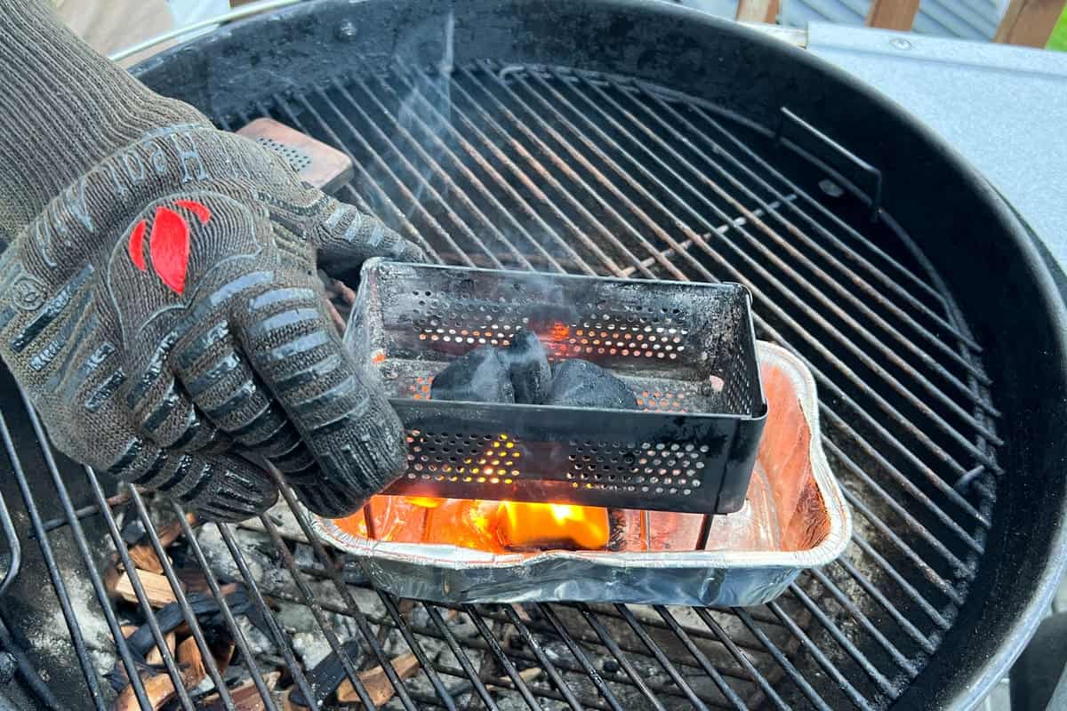 gloved hand placing the smoker box filled with charcoal briquettes into an aluminum foil loaf pan with a lit fire starter inside