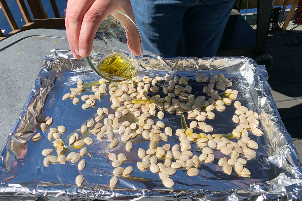 a hand pouring a small glass bowl of olive oil on top of washed pumpkin seeds on a foil lined baking sheet