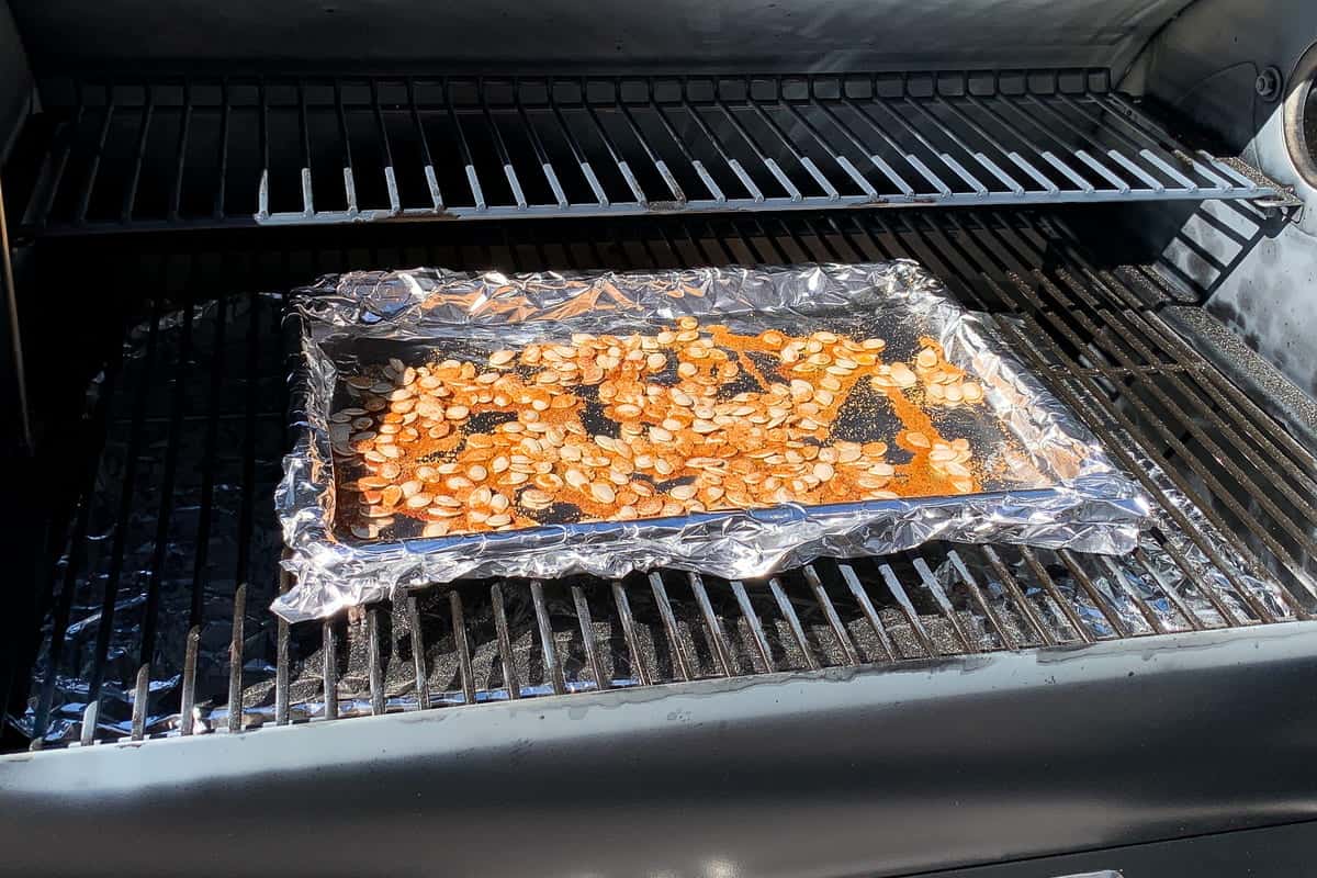 seasoned pumpkin seeds on a foiled lined baking sheet on top of the grill grates of a pellet grill