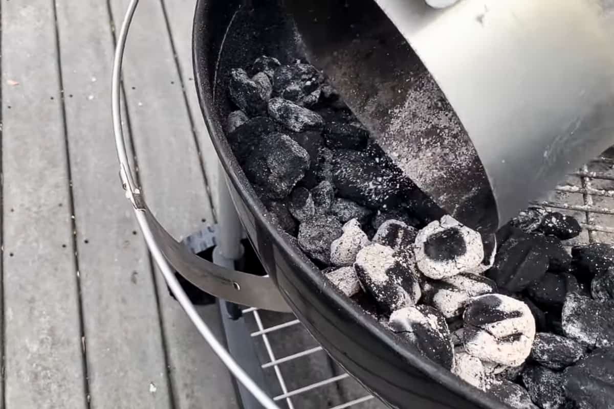 a charcoal chimney filled with lit charcoal being dumped onto a pile of unlit charcoal briquettes