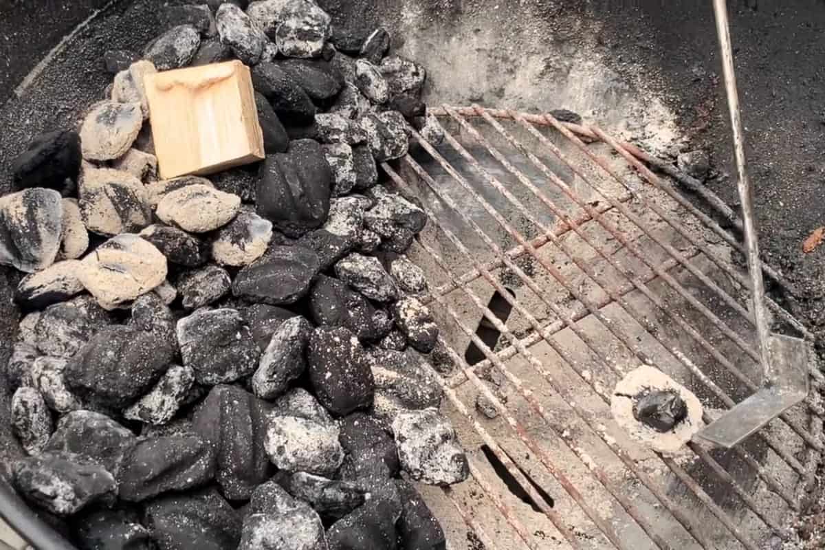 a charcoal briquette on the grill with a plie of briquettes on the other side and a chunk of wood on top