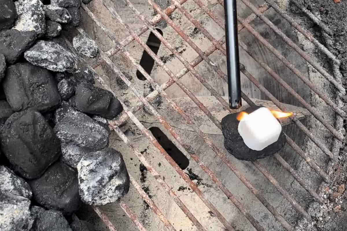 a charcoal grill with a charcoal briquette on one side and a wax lighter cube on top being lit with a wand lighter