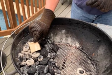 a gloved hand placing a chunk of wood onto a pile of lit charcoal on a charcoal grill