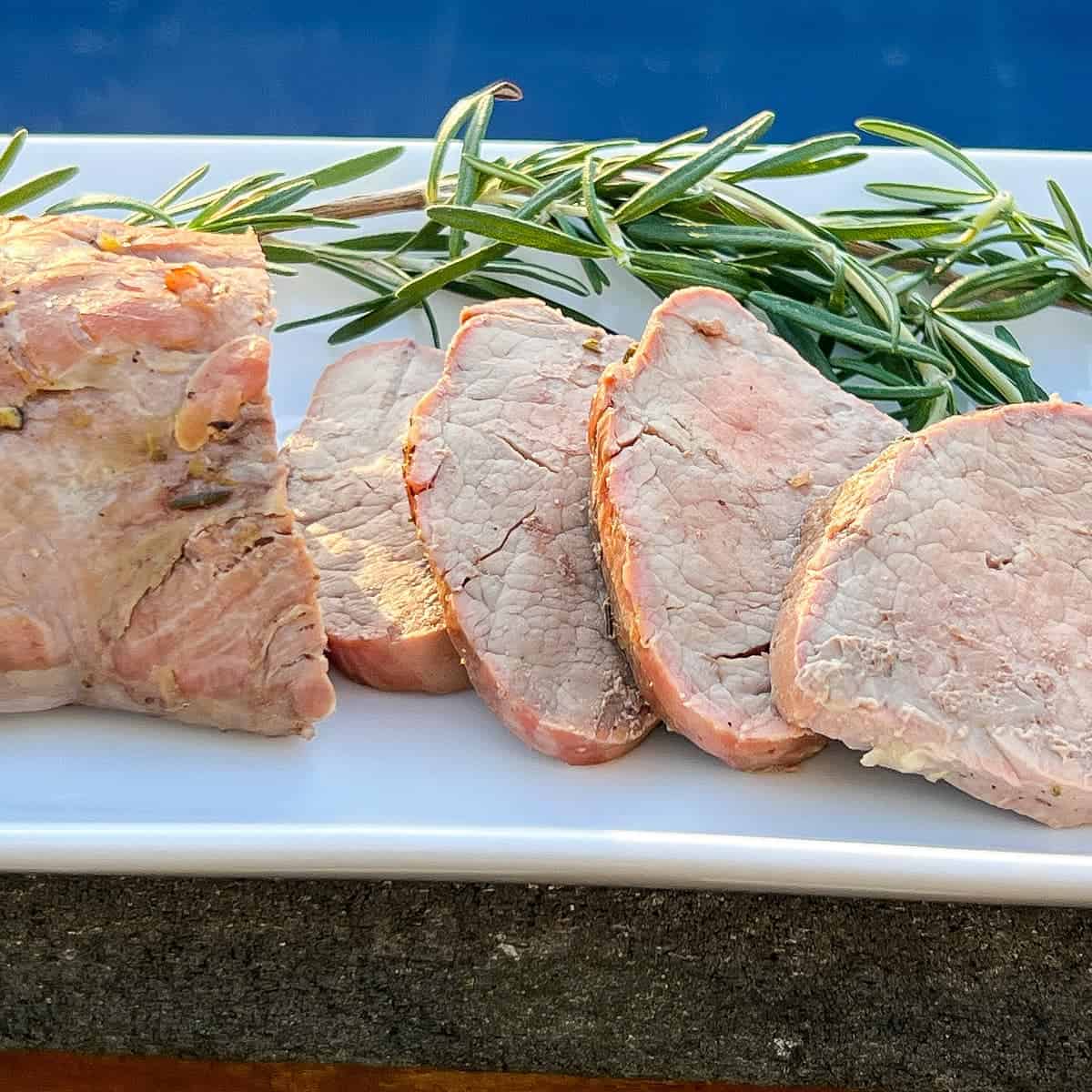 Smoked Pork Tenderloin sliced on a white plate with fresh rosemary on the side as garnish