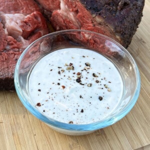 horseradish cream sauce in a glass bowl on a cutting board with smoked beef behind it