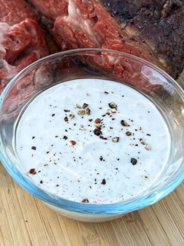 horseradish cream sauce in a glass bowl on a cutting board with smoked beef behind it