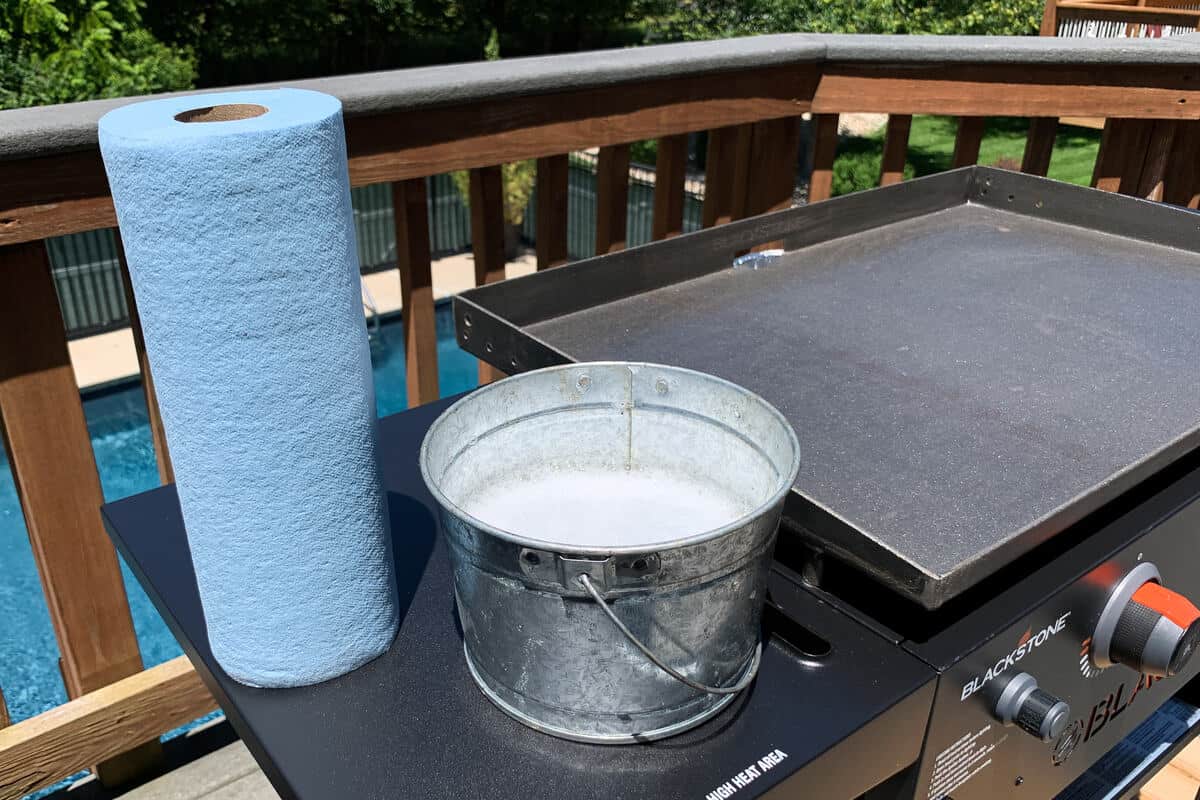 a roll of blue shop towels and a bucket of soapy water on the side shelf of Blackstone griddle