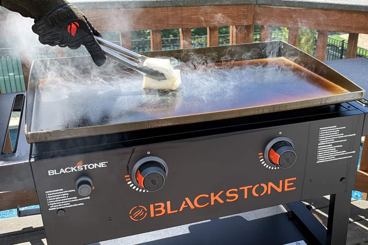 a paper towel held with BBQ tongs pushing around seasoning oil across the surface of a Blackstone griddle