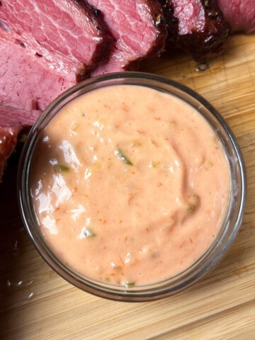 thousand island dressing in a glass bowl on top of a wooden cutting board next to slices of smoked corned beef