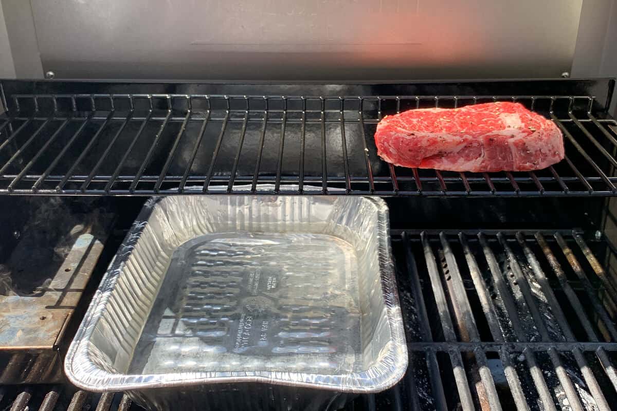 a ribeye steak on the top rack of gas grill with a water pan and smoker box on the bottom grates