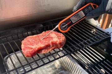 a steak on the top rack of a gas grill with an instant read thermometer measuring 90 degrees internally