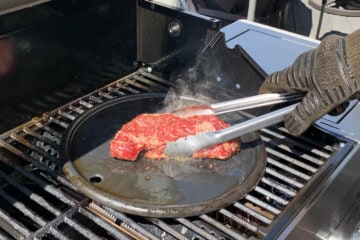 a ribeye steak searing on a cast iron pan with a pair of metal tongs moving it around