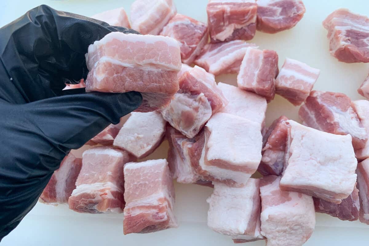 a gloved hand holding up a cube of raw pork belly