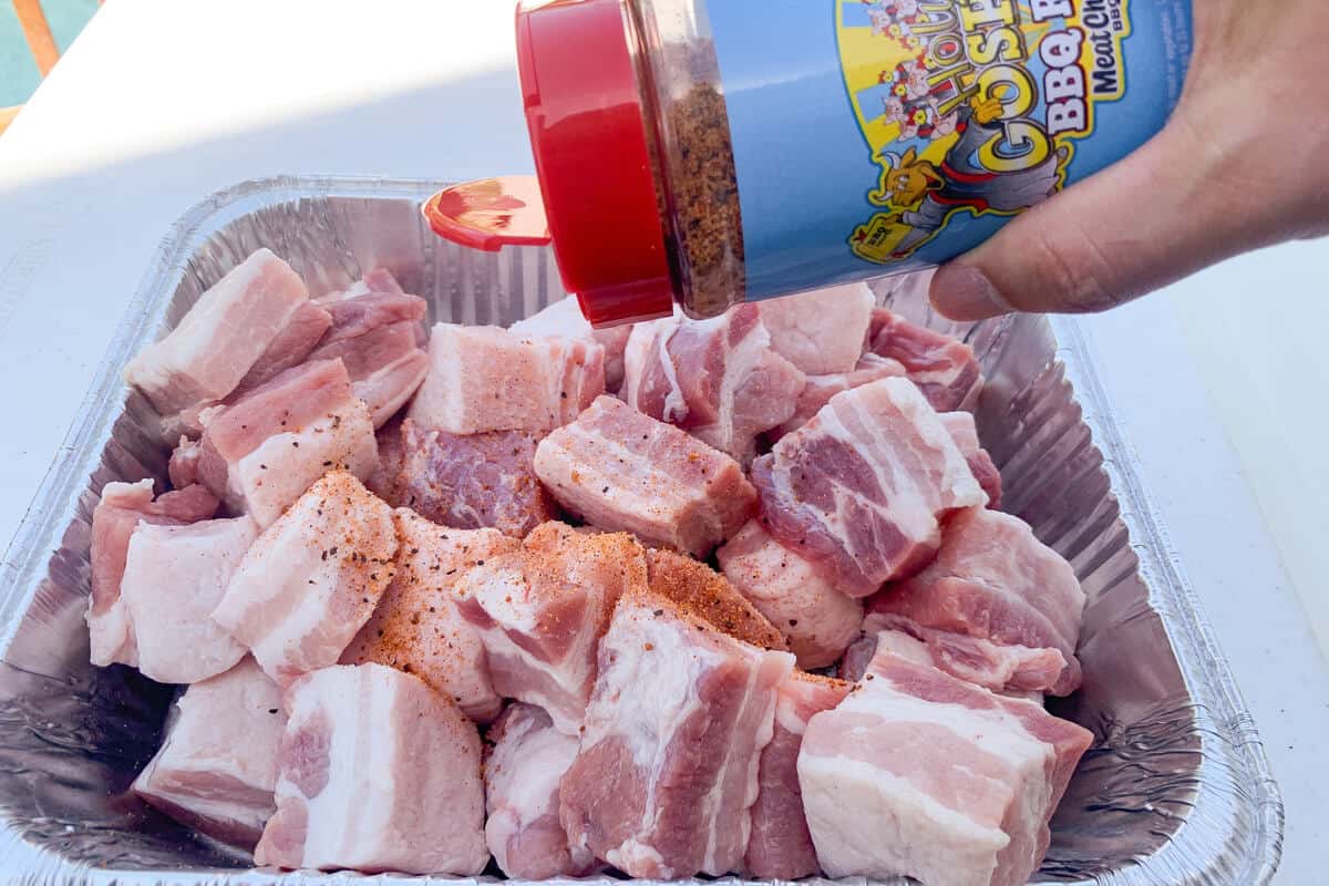 Meat Church The Gospel rub being sprinkled over chunks of raw pork belly