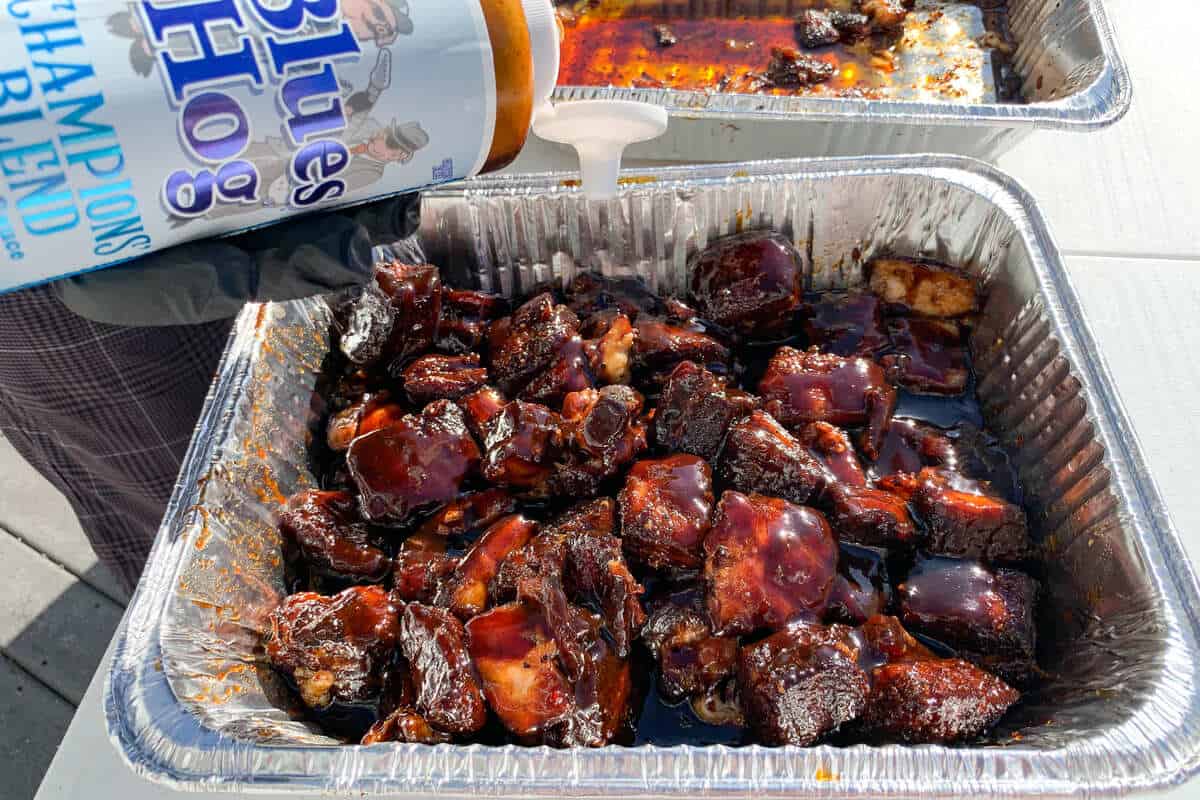 pork belly chunks in a foil pan being drizzled with BBQ sauce