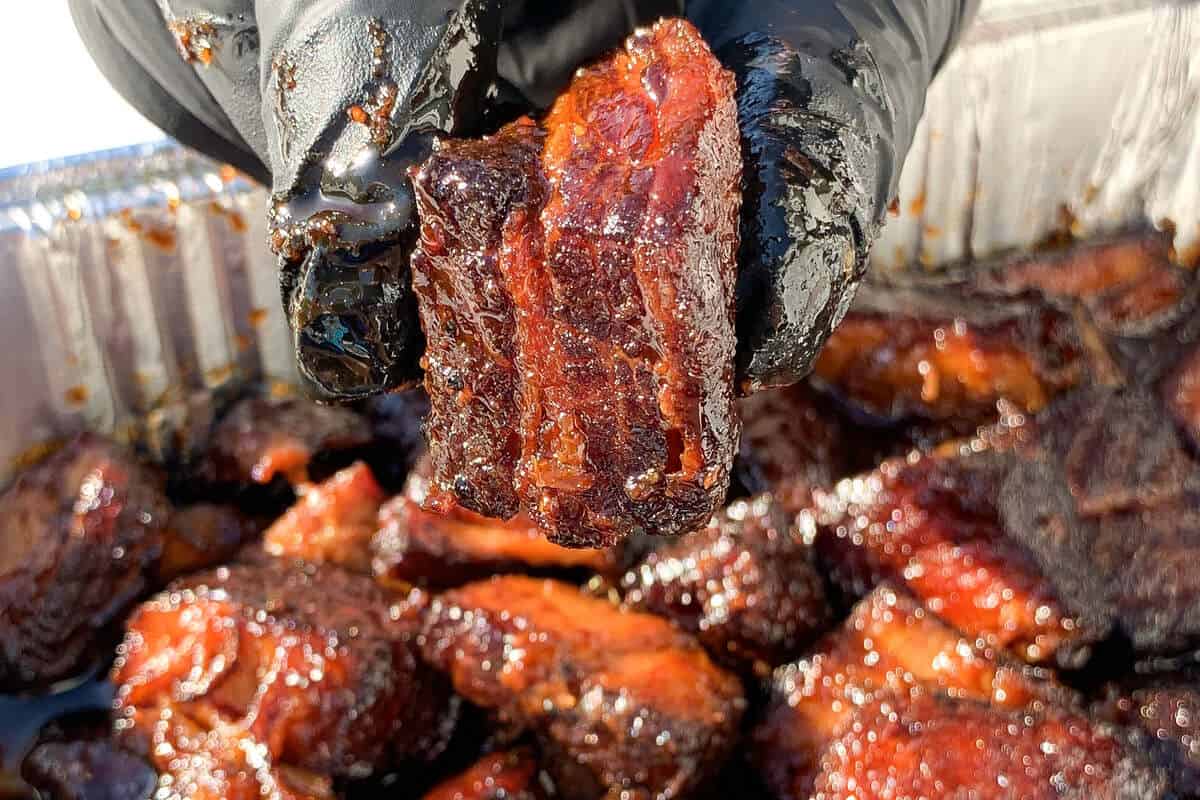 a gloved hand holding up a chunk of pork belly burnt ends