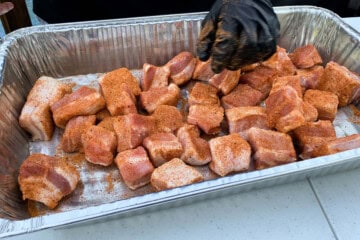 cubes of pork belly in a foil pan being seasoned with a gloved hand