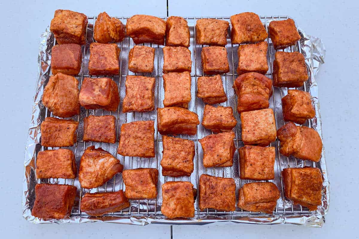seasoned cubes of raw pork belly on a wire rack set over an aluminum foil lined baking sheet
