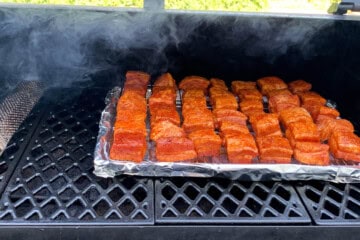 a pan of seasoned pork belly cubes on the grates of a pellet grill with a smoker tube next to it