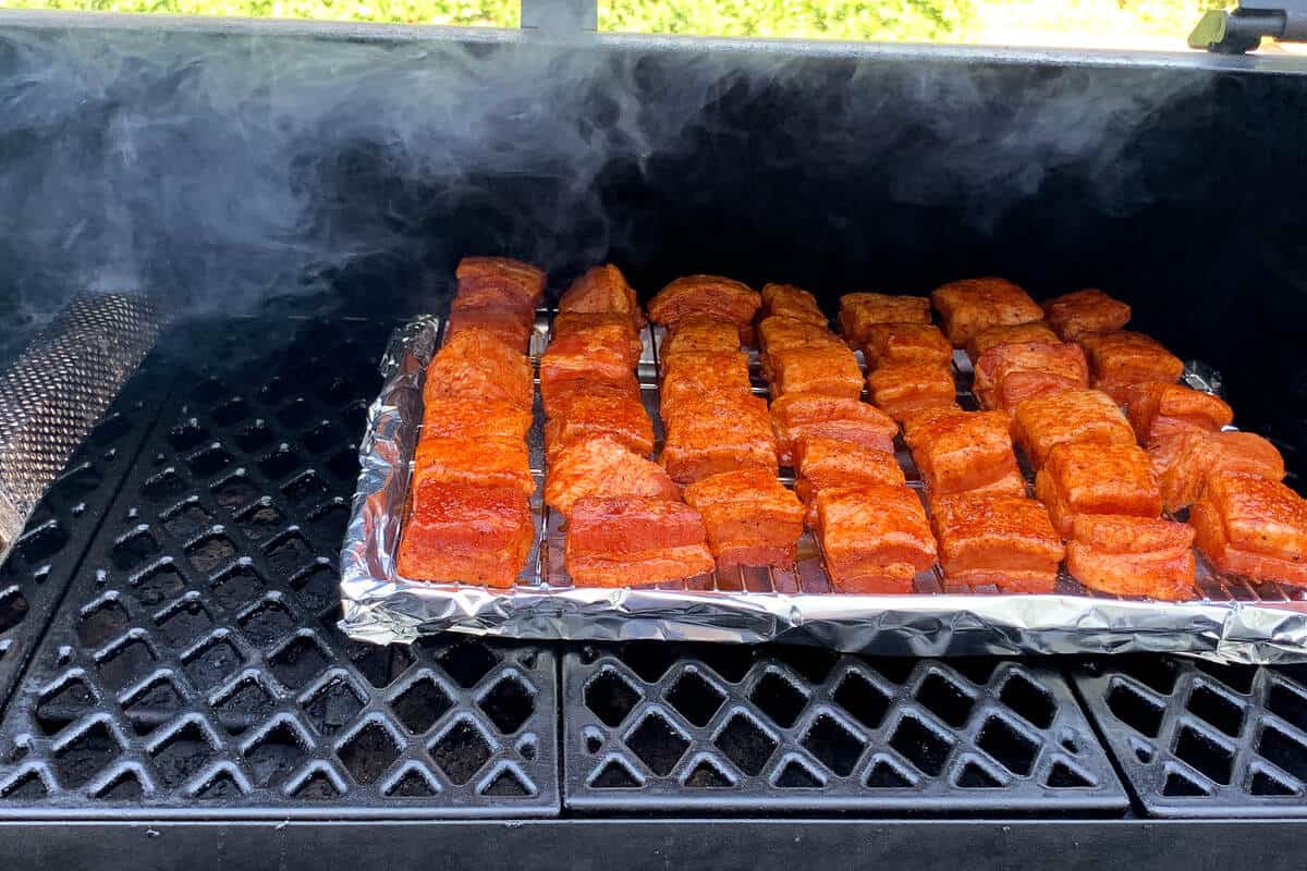 a pan of seasoned pork belly cubes on the grates of a pellet grill with a smoker tube next to it