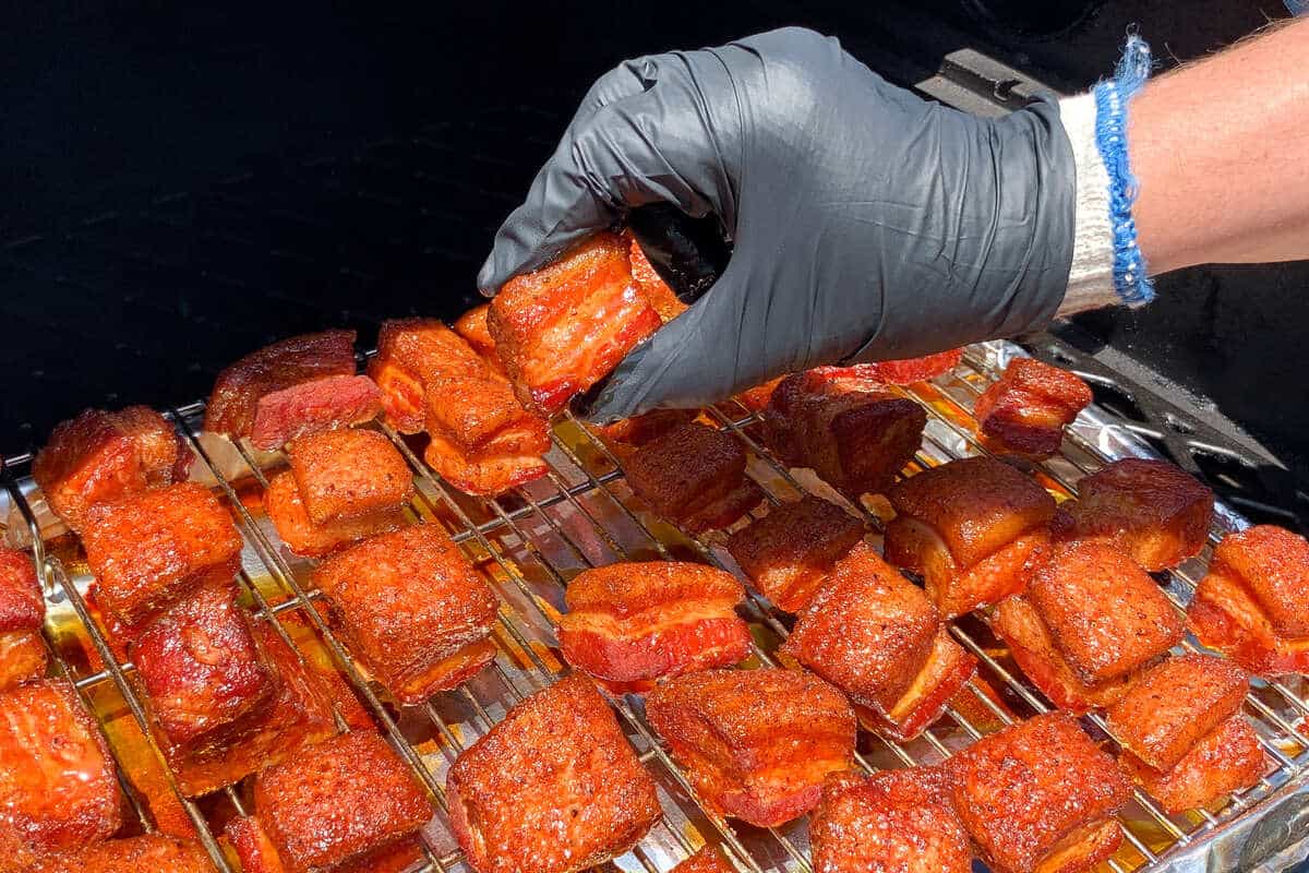 a gloved hand holding up a cube of seasoned pork belly to show that the fat has not yet completely rendered