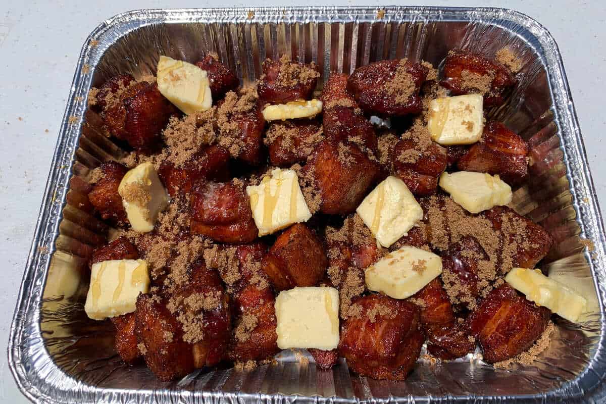 an aluminum foil pan filled with seasoned pork belly cubes with brown sugar sprinkled on top, slices of butter, and honey drizzled