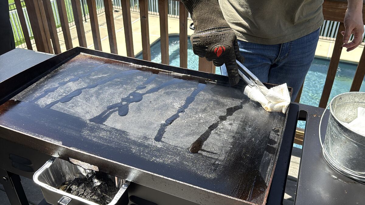 oil drizzled on the surface of a Blackstone griddle with paper towels in tongs spreading the oil