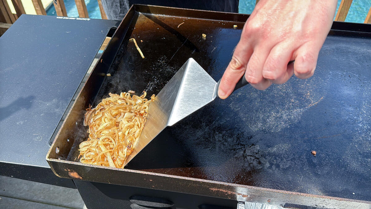 grilled onions being pushed to the side of the Blackstone griddle with a metal spatula